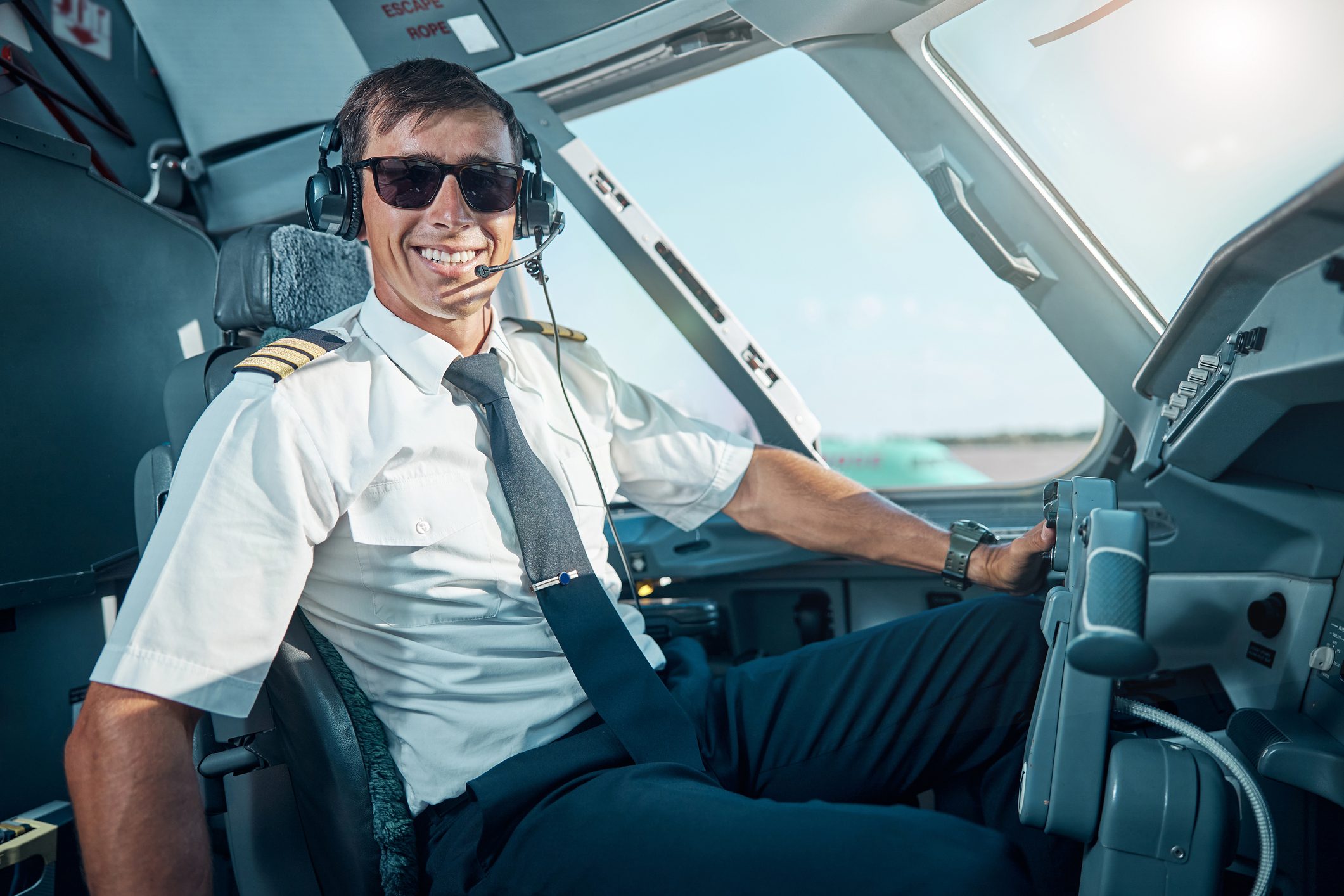Waist up portrait of cheerful young man in glasses and earphones sitting in cockpit and getting ready for flying